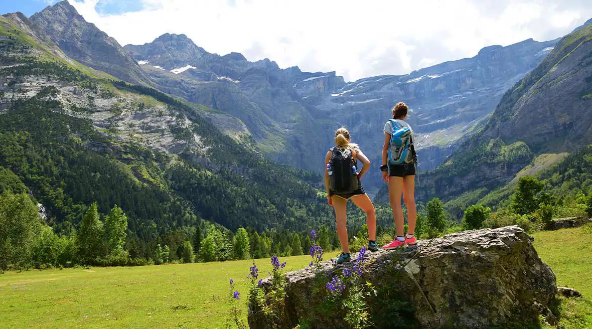 Two tourists standing on a rock in the middle of a grassy field staring at the mountains beneath a blue sky with white clouds and green trees at the base of the mountains