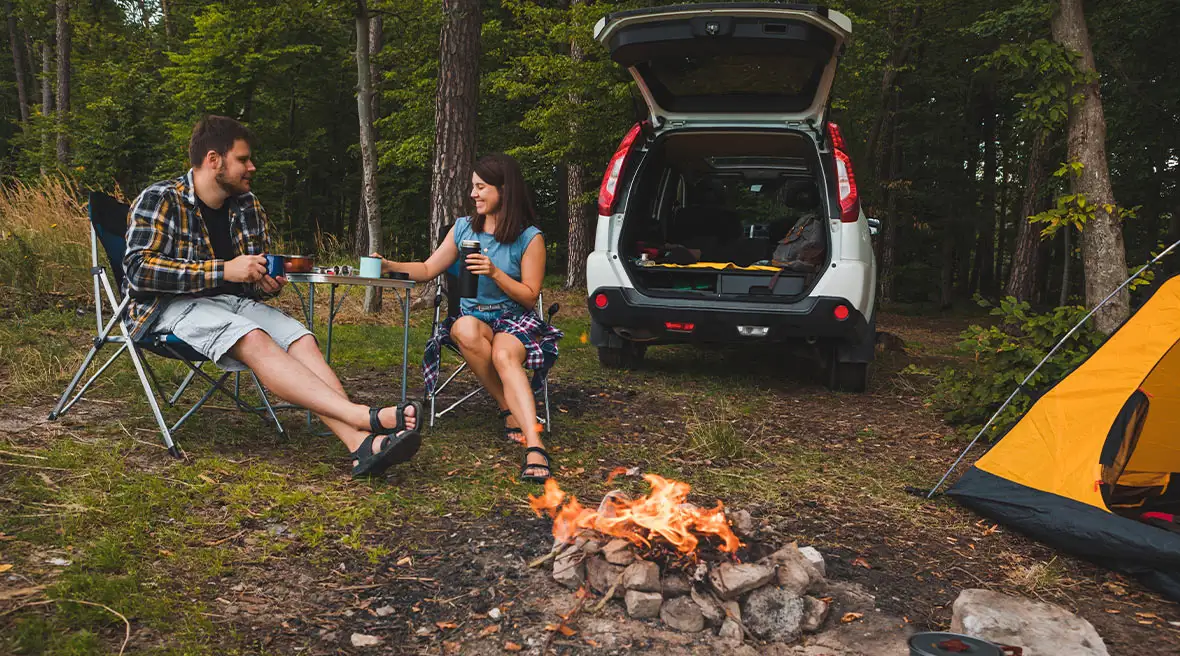 Man and woman sitting on the grass by their car and their yellow tent by a fire