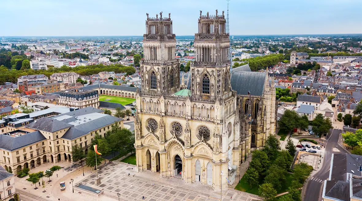 An imposing cathedral with two towers stands high above a city square, surrounding buildings and green spaces