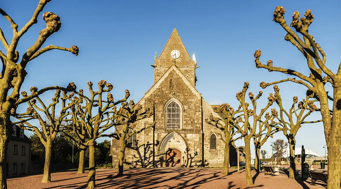 An old stone-built church in the background with a white parachute just visible, caught up on part of the roof. Trees and their shadows dot the foreground.