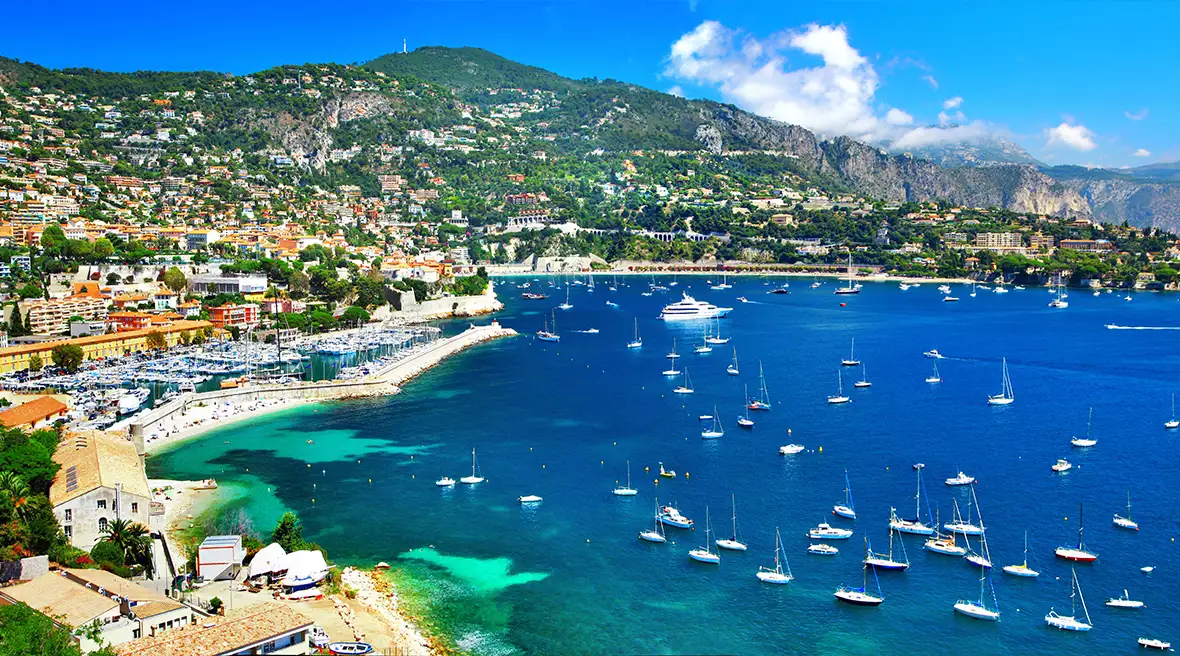 Clear blue water in a bay with boats and buildings across hills in the distance