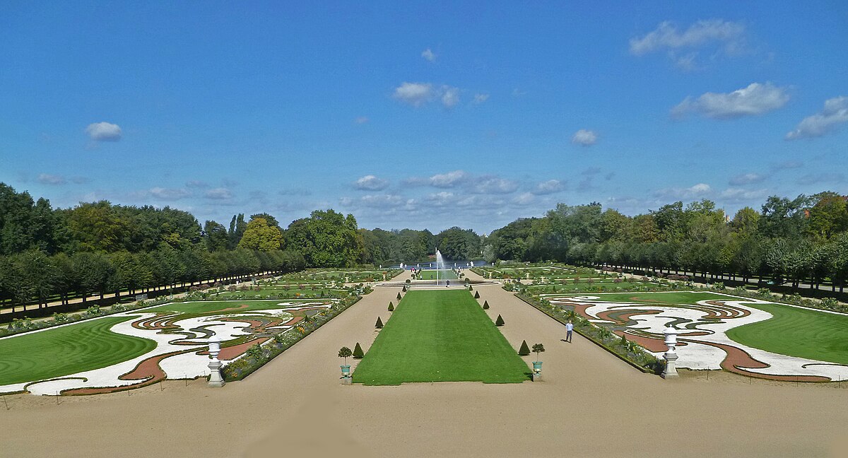 A view of the Charlottenburg Palace Gardens. Symmetrical and well-manicured lawns frame two paths. Trees line the outer perimeter and a fountain sits in the centre.