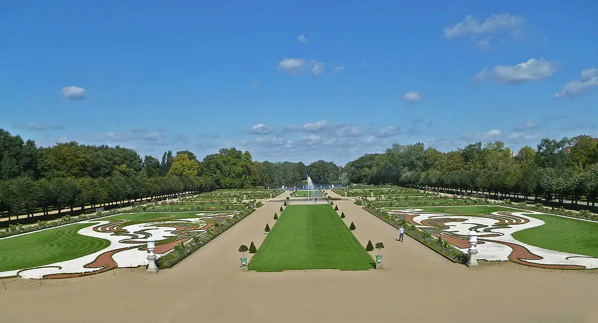 A view of the Charlottenburg Palace Gardens. Symmetrical and well-manicured lawns frame two paths. Trees line the outer perimeter and a fountain sits in the centre.