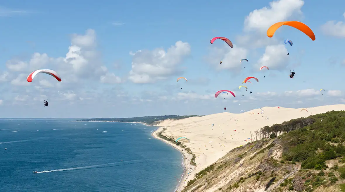 Numerous colourful paragliders sailing out over a huge sand dune and blue waters on a backdrop of blue sky with fluffy white clouds
