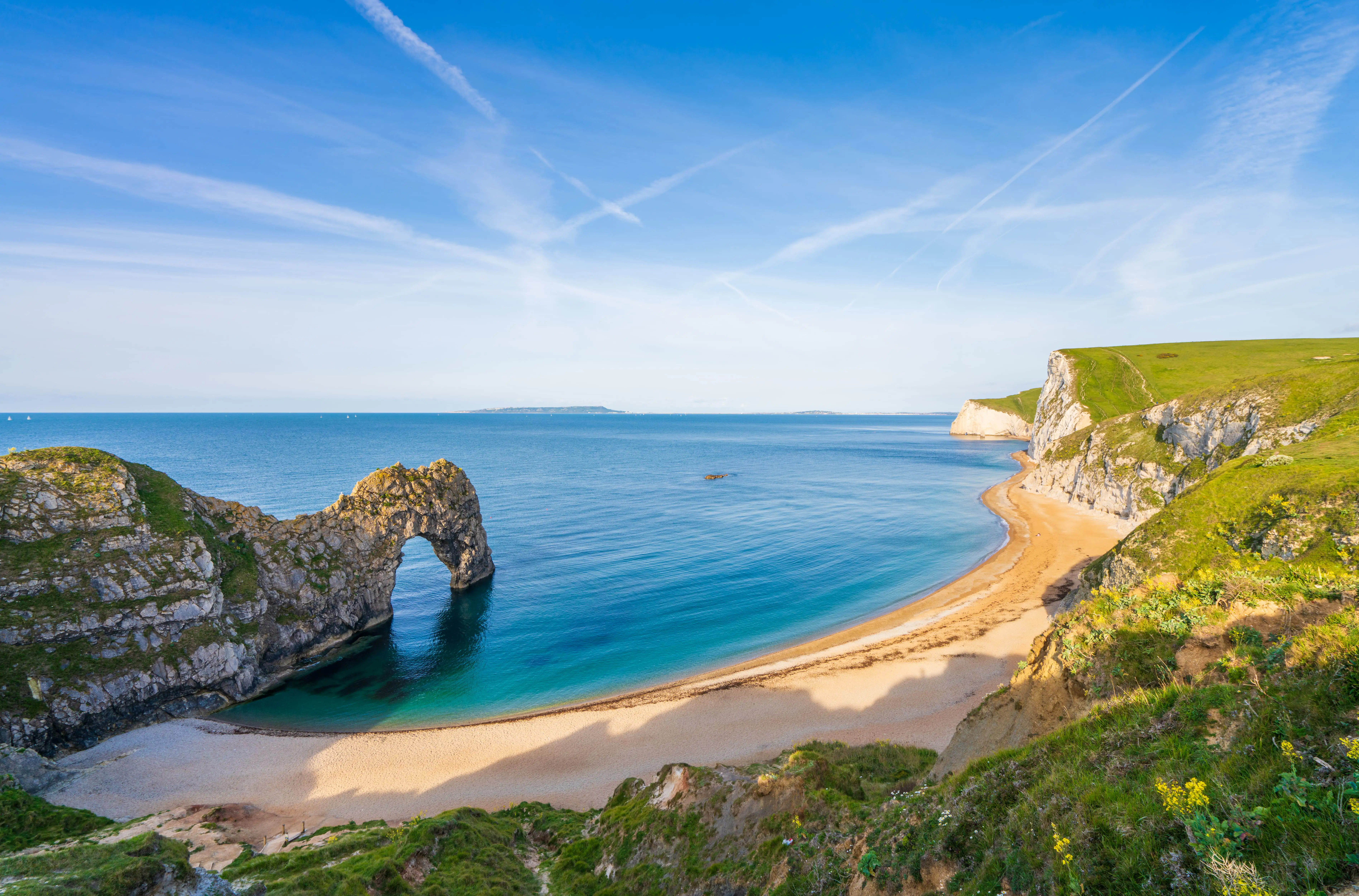 Arche de Durdle Door dans la mer bleu clair, avec une plage de sable doré, des falaises herbeuses et un ciel bleu.