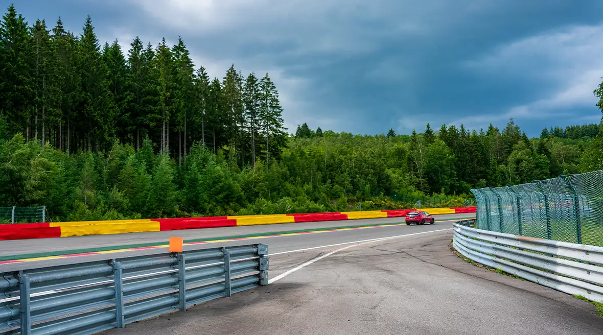 Red car driving round a bend on the Circuit de Spa-Francorchamps, with crash barriers either side of the track and forest outside the track.