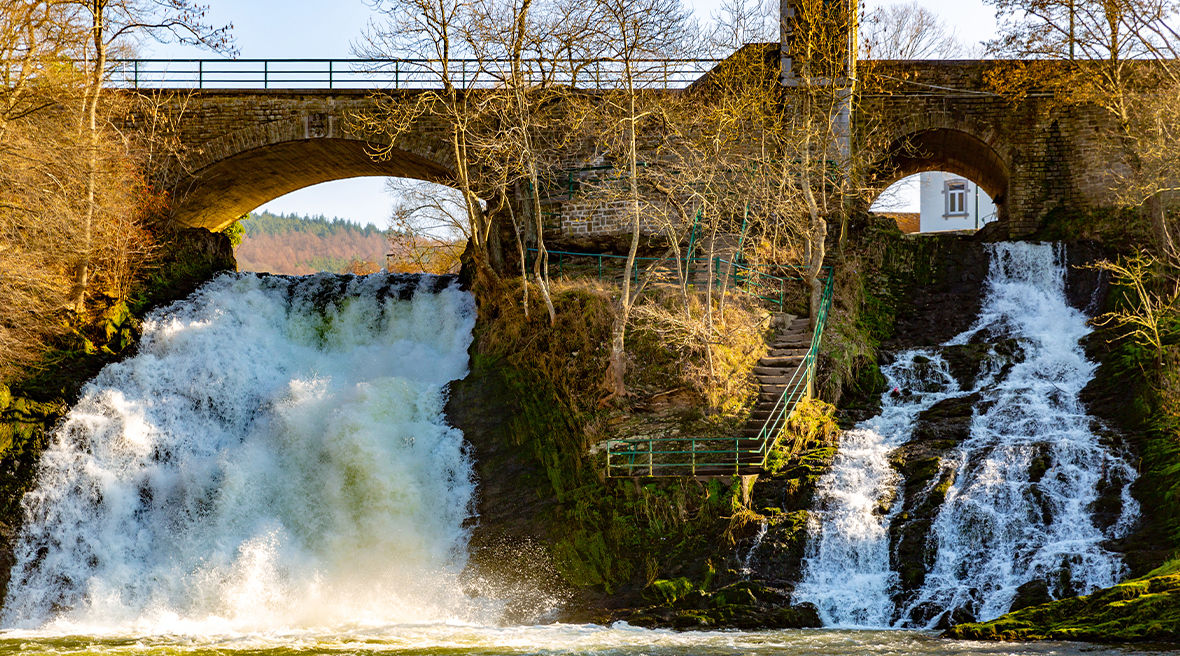 The Coo waterfall, near Stavelot, flowing under a stone road bridge surrounded by leafless trees on a sunny winter’s day.