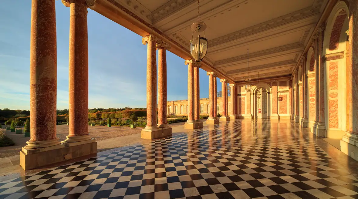 A colonnade of a grand building, with pink classical columns, arches and a black and white chequerboard floor
