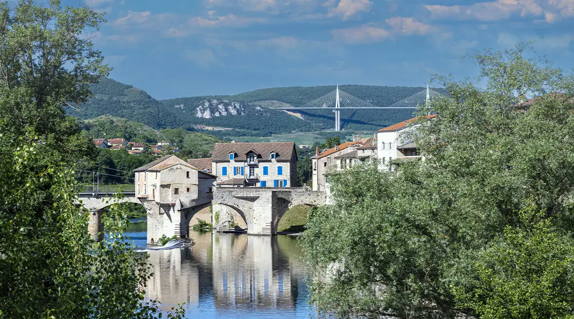 A massive road bridge can be seen in the distance from a viewpoint over a river with an old bridge in the foreground