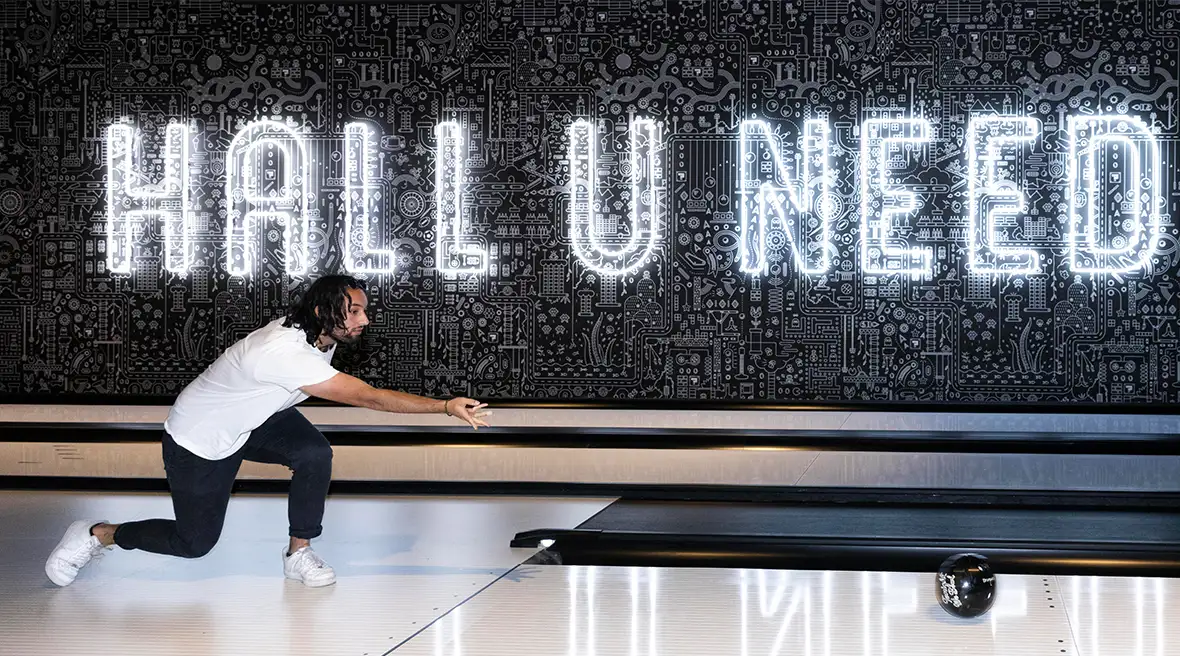A side view of a young man throwing a ten-pin bowling down an alley with the words Hall U Need lit up behind him