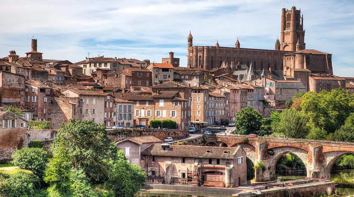 A city filled with many stone Gothic buildings with an arched bridge over a river in the foreground and a large church in the background