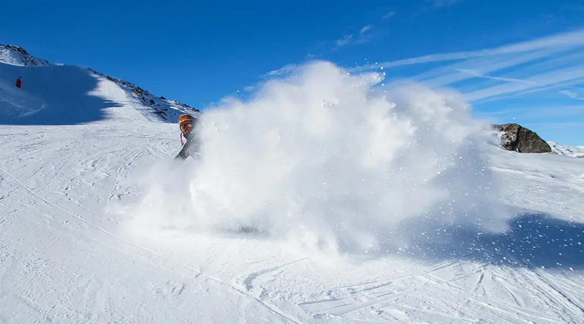 snowboarder in a woolly hat creating a huge billow of snow as he slides down the side of a mountain