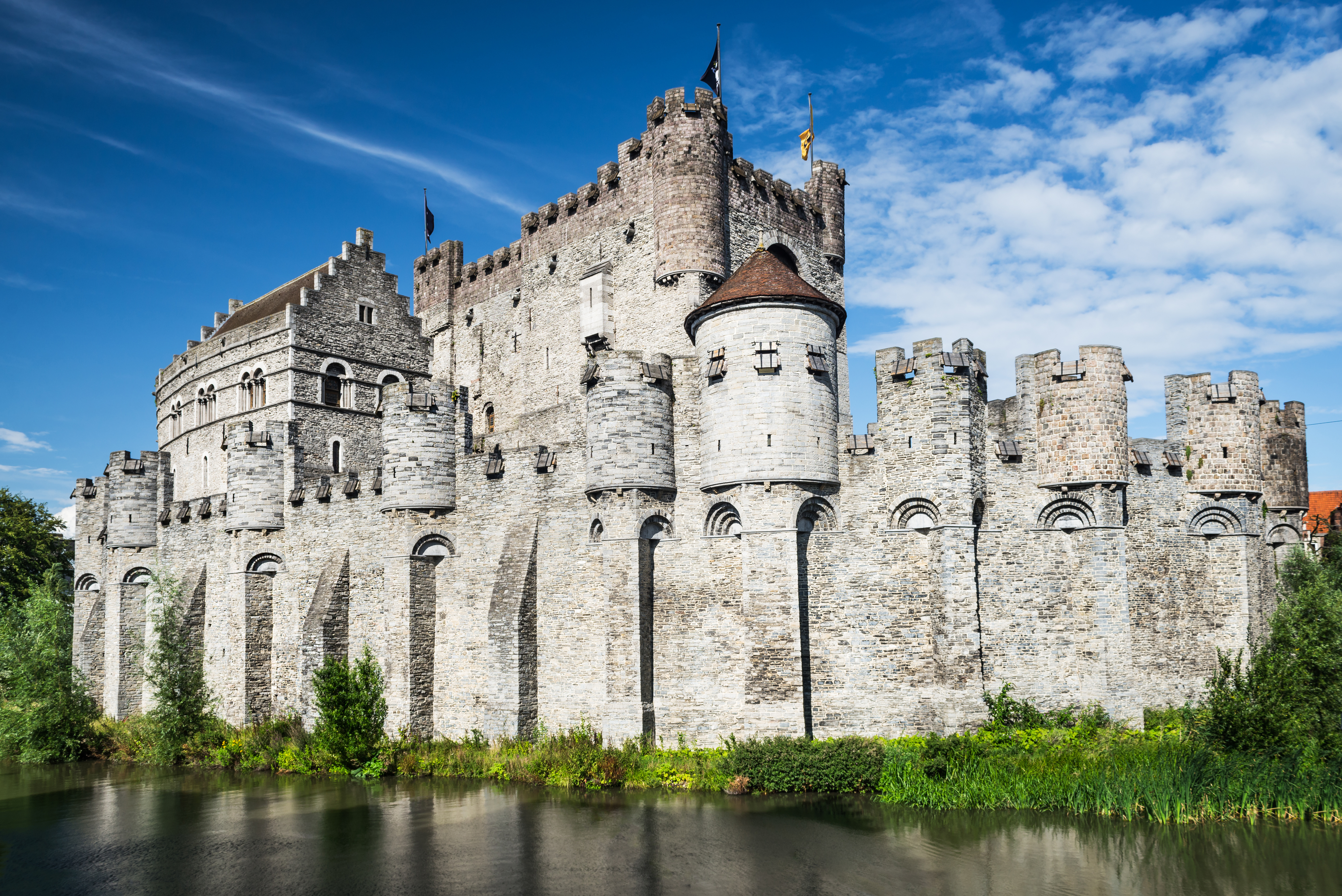 Gravensteen Castle, an imposing building with military architecture overlooking the Lieve River, against the backdrop of a blue sky.