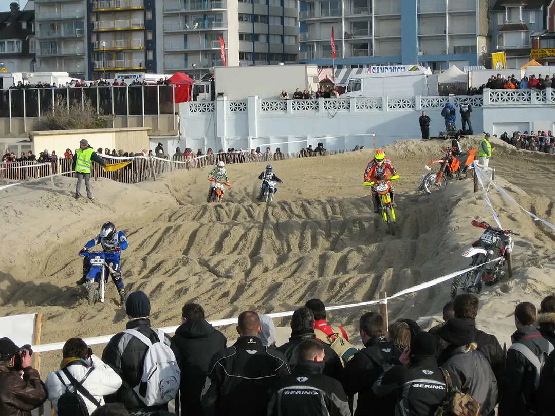 A group of motorbikes racing at the Enduropale du Touquet, a competition which takes place every February, on sand mounds, with a few spectators watching and stewards.