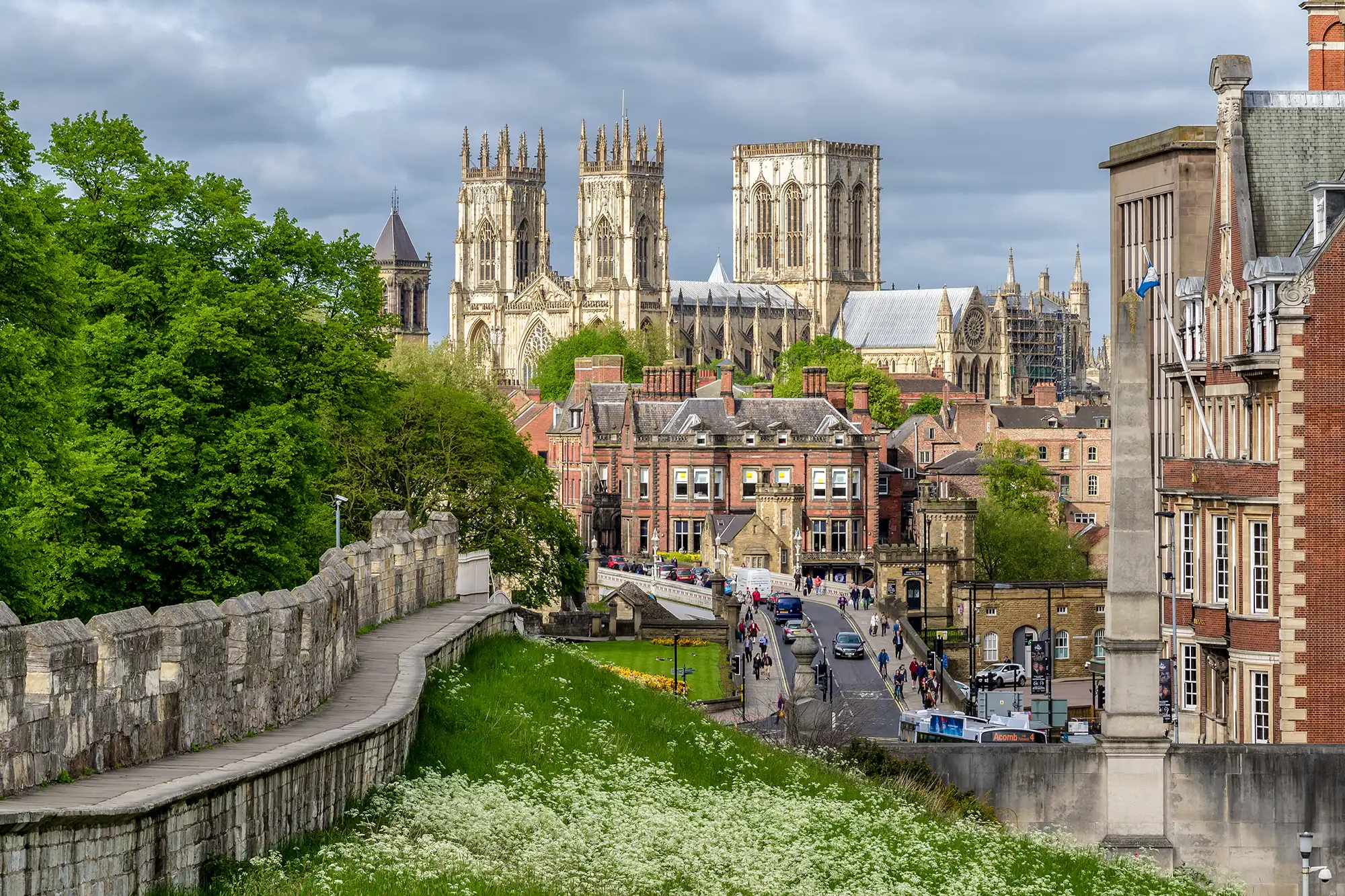 Une vue de la ville de York avec ses murailles sur la gauche, une route avec des piétons bordée de grands bâtiments en briques et York Minster en arrière-plan qui s’impose sur toute la ville