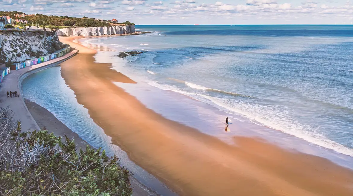 Un surfeur dans l’eau tenant sa planche sur la plage de Stone Bay. Des cabines de plage colorées sont disposées dans le sable au pied de hautes falaises blanches