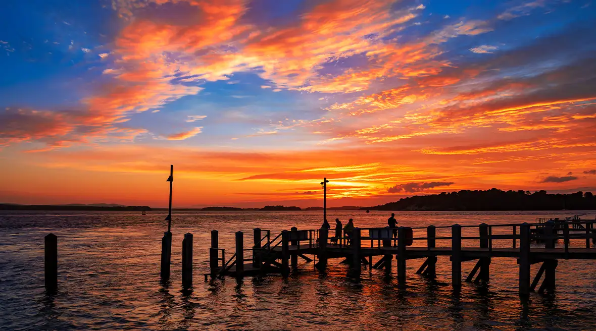 Personnes admirant le coucher de soleil sur un petite jetée en bois à Sandbanks. Le ciel bleu foncé est éclairé par le soleil se reflétant sur les nuages leur donnant des nuances d’orange.