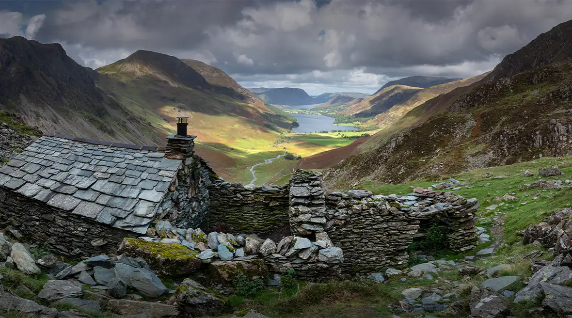 Une vieille maison en pierre et en ardoise au-dessus d’une vallée verdoyante menant à un lac entouré de montagnes, sous un ciel orageux