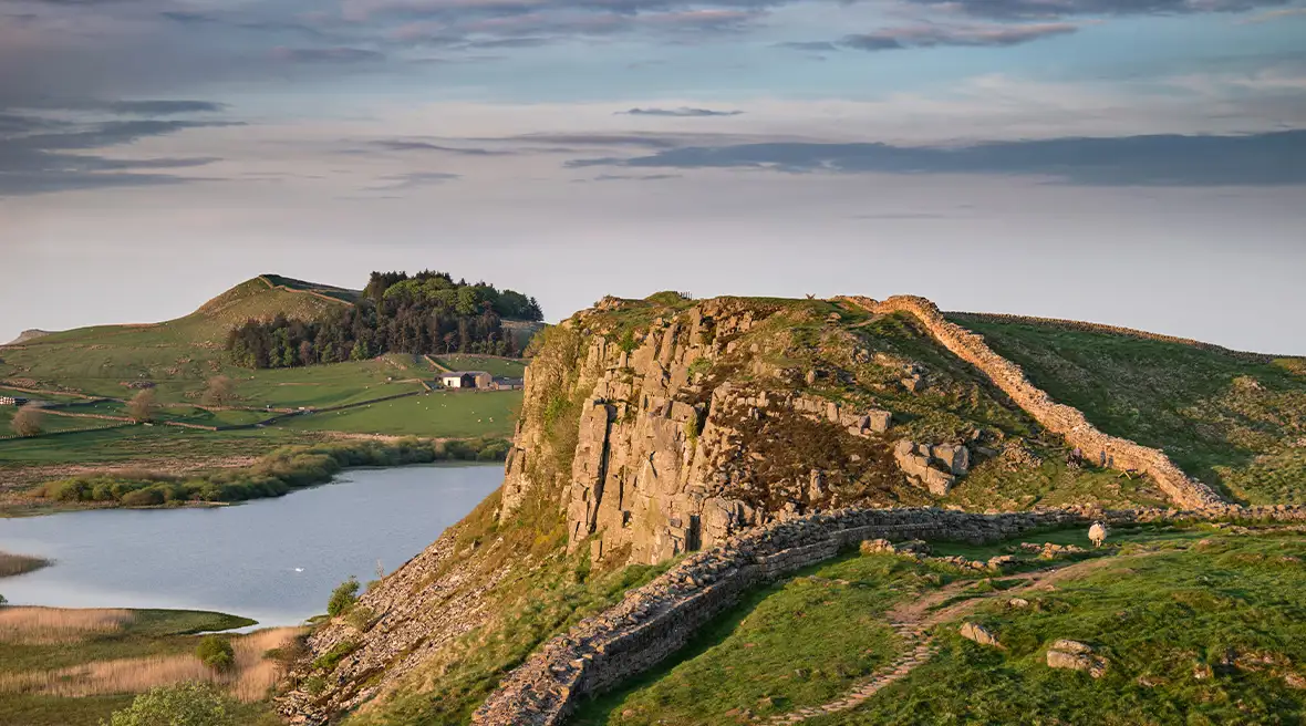 Le mur d’Hadrien en pierres s’étend sur une falaise verte dominant la mer avec une colline, des arbres et une colline en arrière plan