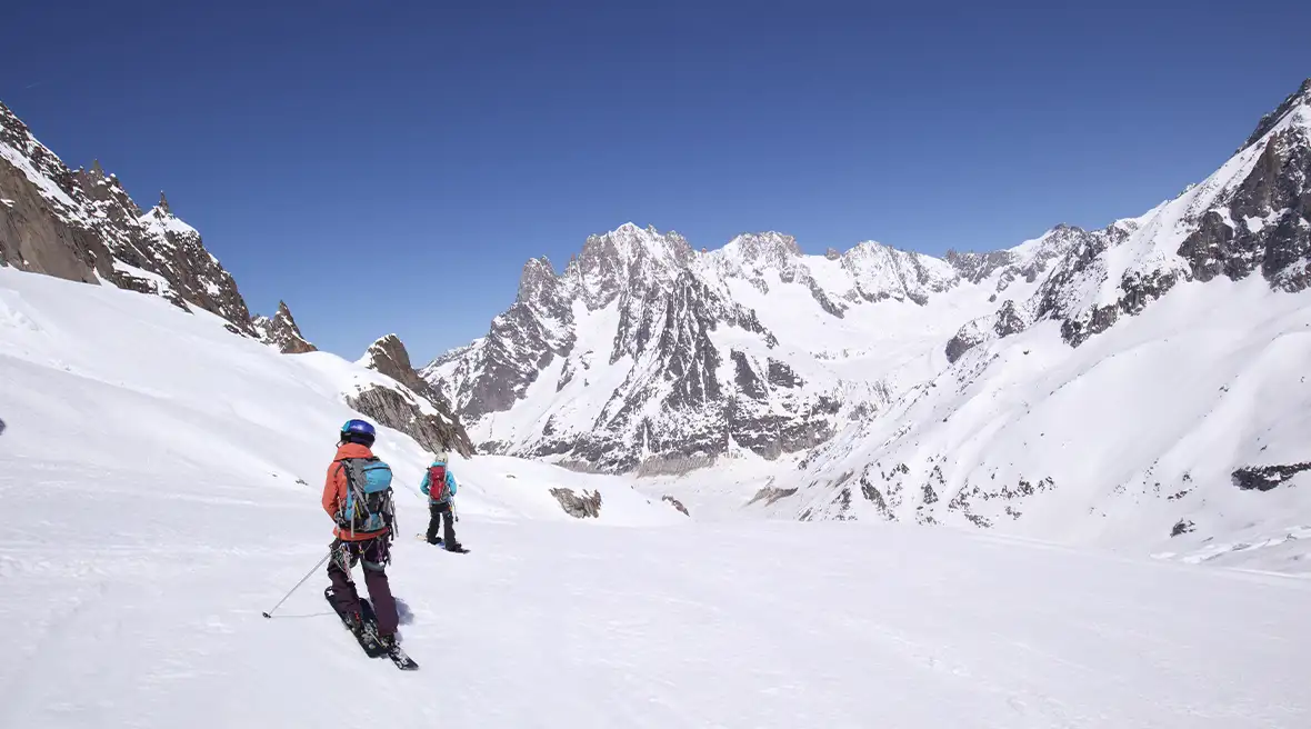 Two skiers skiing down a wide slope with tall snow-capped peaks in the distance