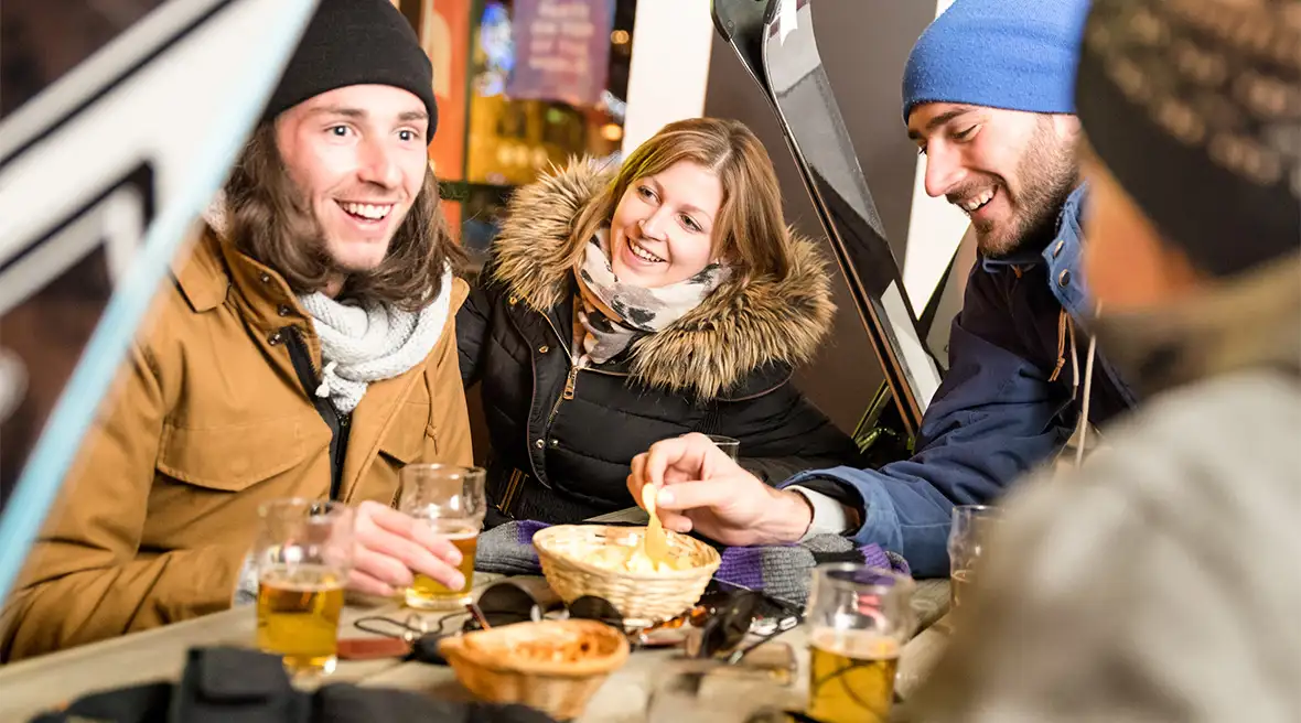 A group of friends drinking beer and eating crisps with skis in the backround