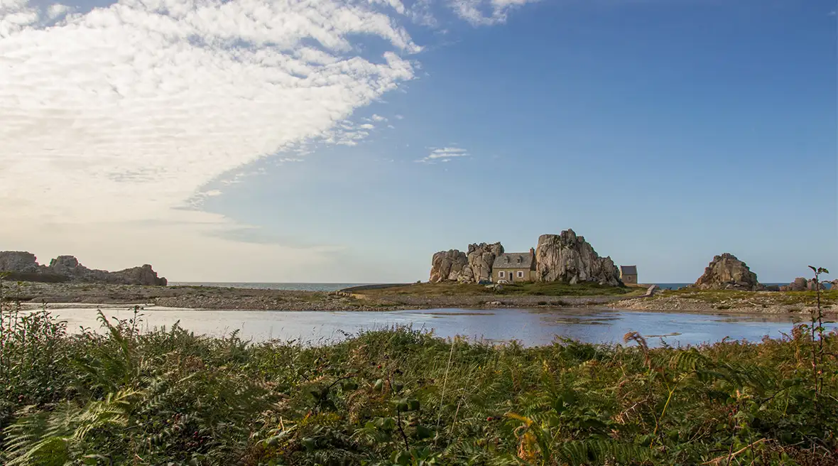 A tiny stone-built cottage sits nestled between two huge rocks on the seashore