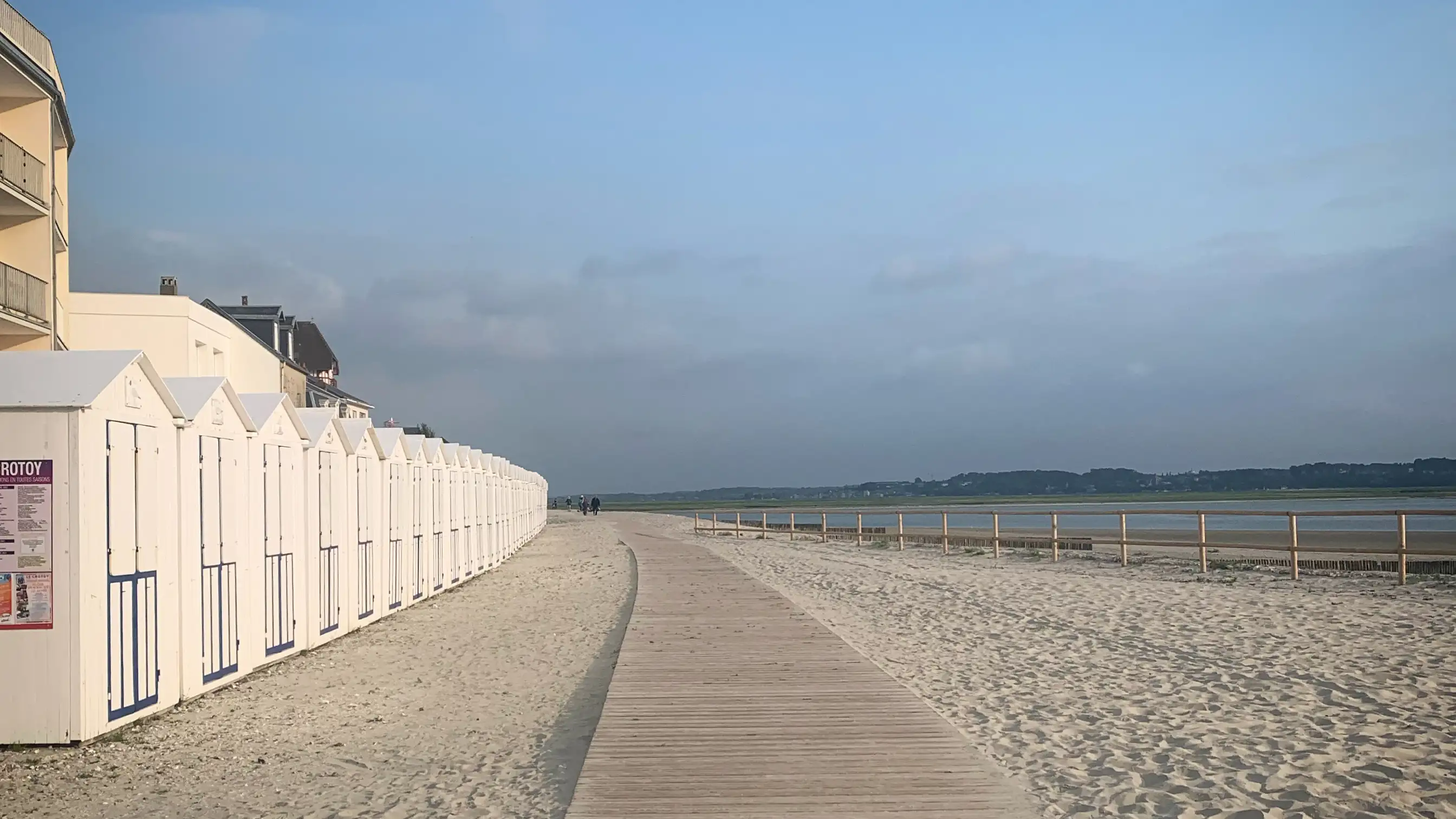 A woodend slatted footpath and a row of white beach huts on the white sands of the bay of the Somme