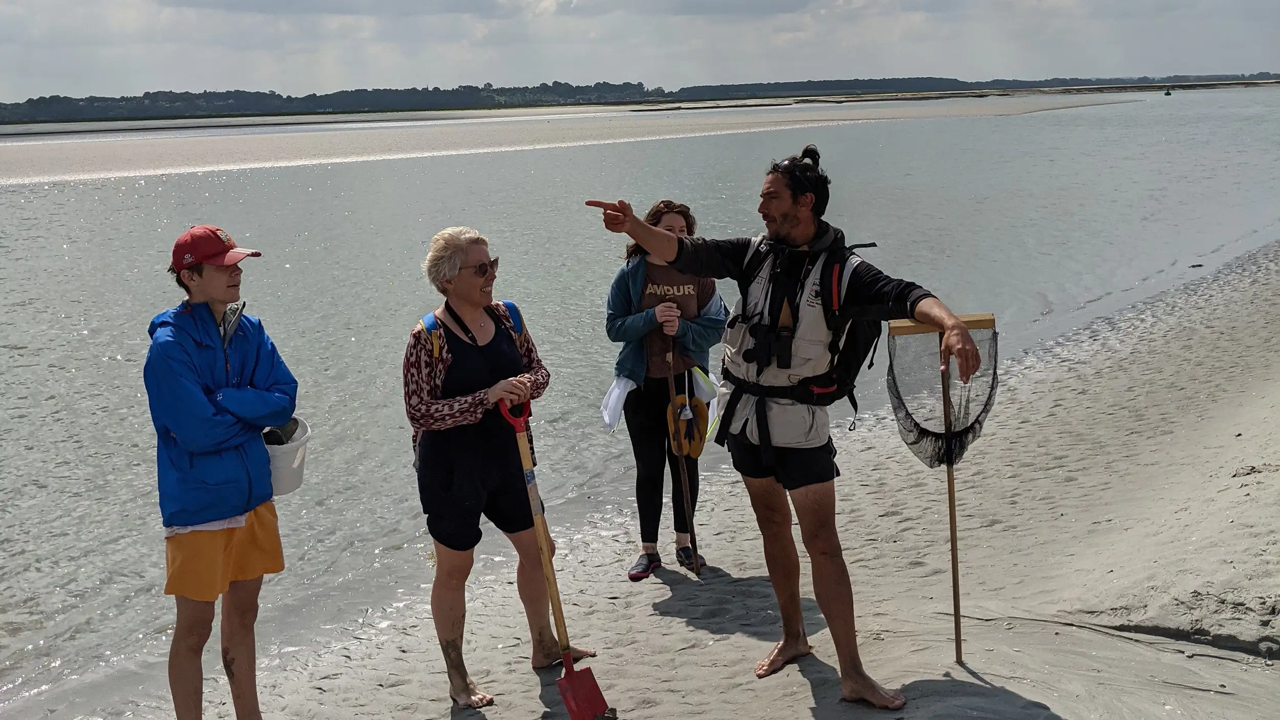 A male tour guide holding a big wooden fishing net tells a group of three tourists about the wildlife in the sandy bay
