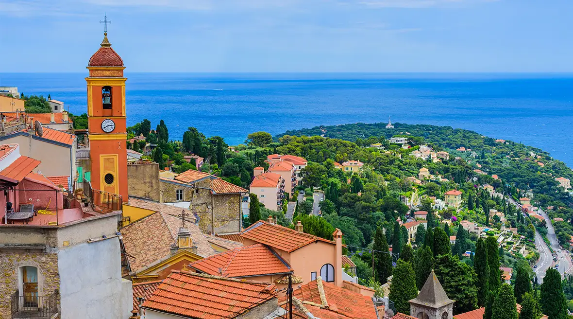 A group of red tile roofed buildings atop a hill overlooking the villages below and the sea in the distance