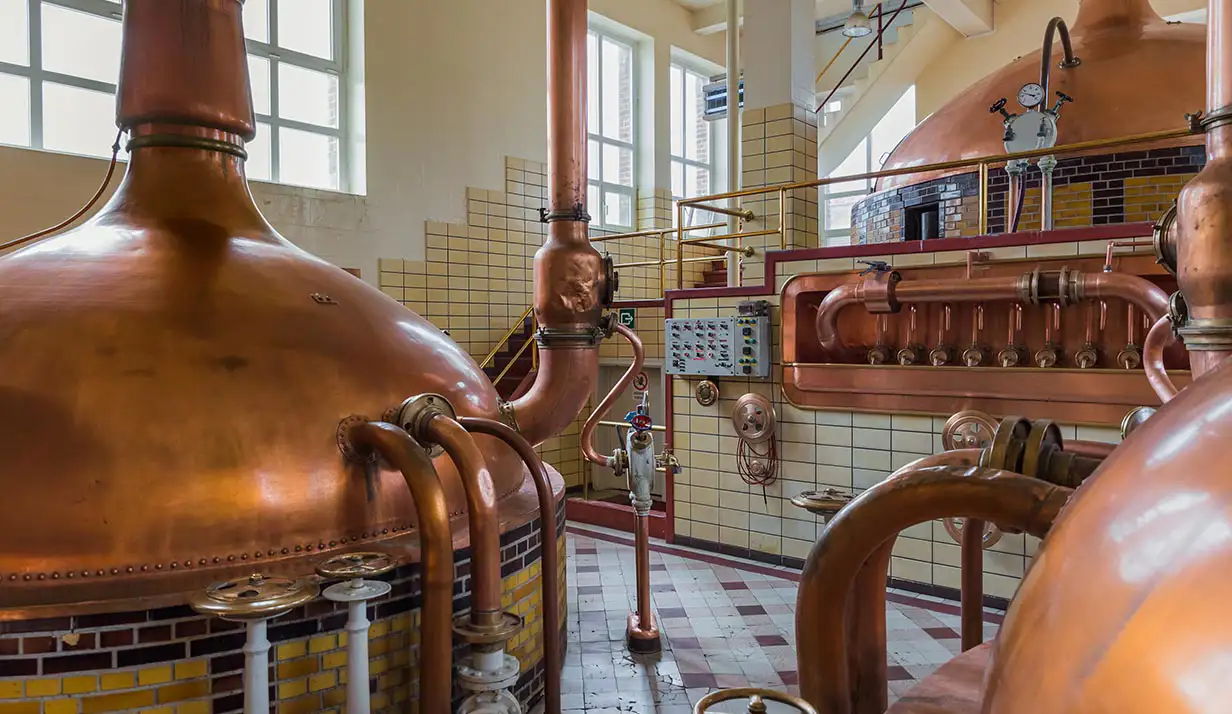 A room with chequered tile flooring containing large copper metal drums for brewing beer inside