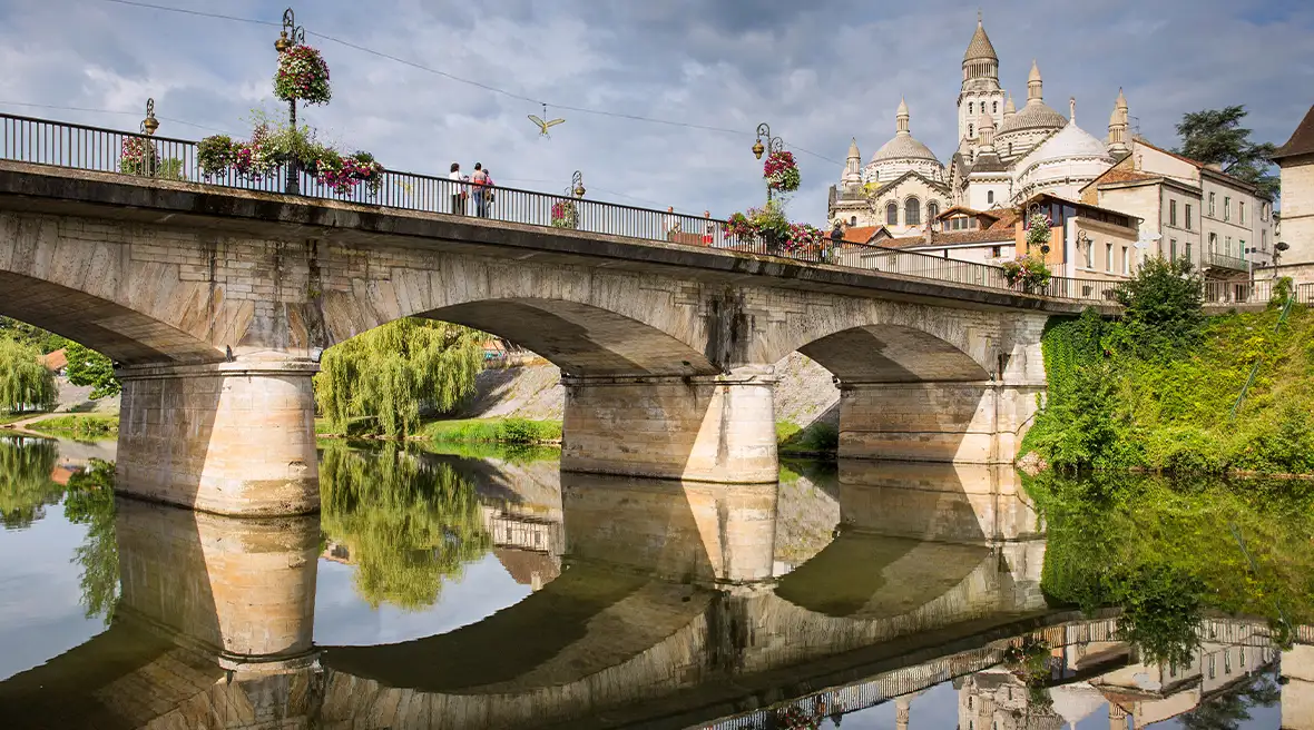Arched bridge over a river with a white stone town to the right with flowers and greenery surrounding it