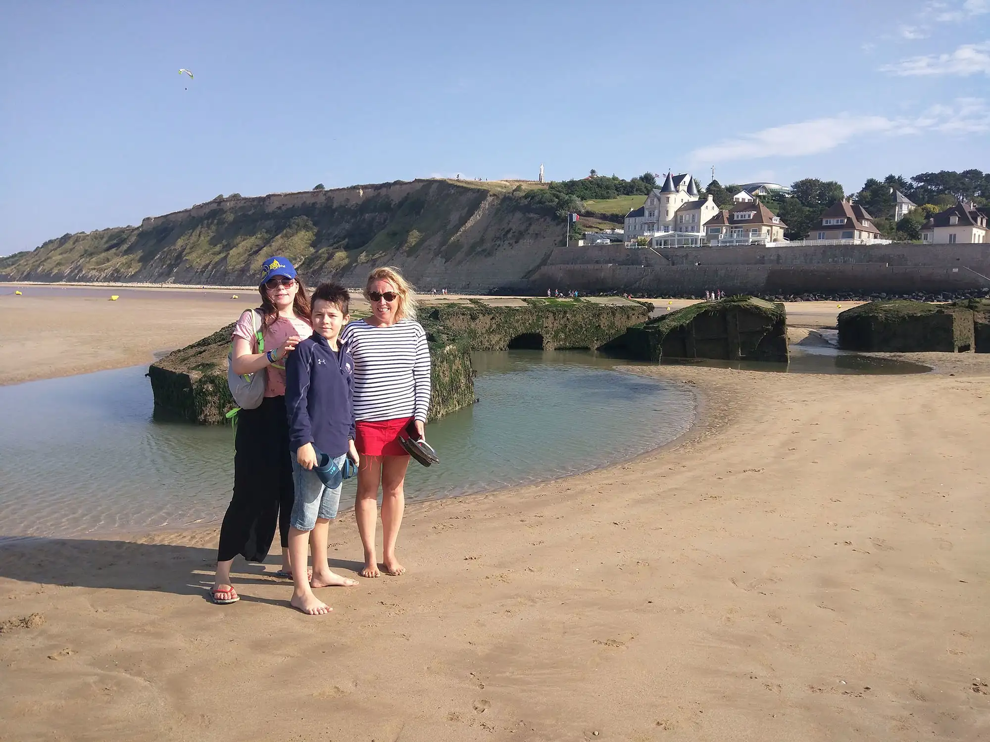 A family of 3 stand next to huge concrete blocks from Mulberry Harbour on the beaches of Normandy.