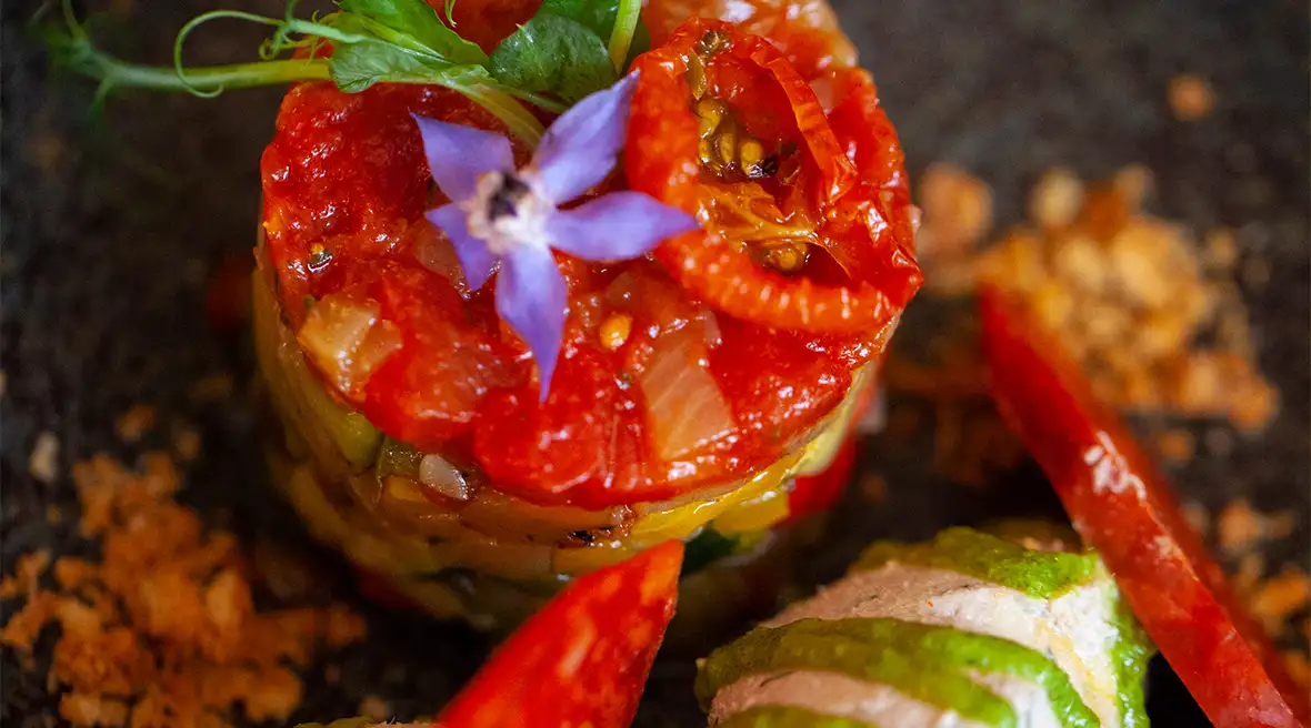 A dark circular plate with a round terrine with a top layer of bright tomato sauce and garnished with a purple flower and delicate leaves. It’s accompanied with two quenelles of pale mousse and zig zags of green dressings on top.