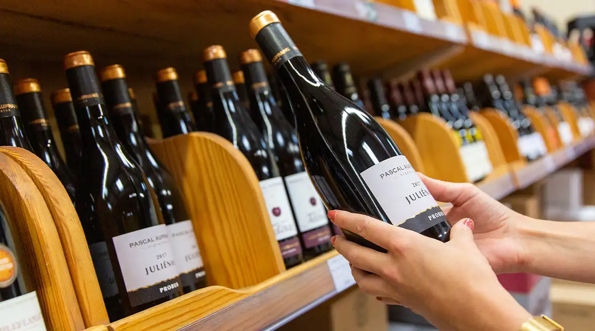 A close up of someone taking a bottle of red wine from a wooden shelf in an aisle in Calais Vins