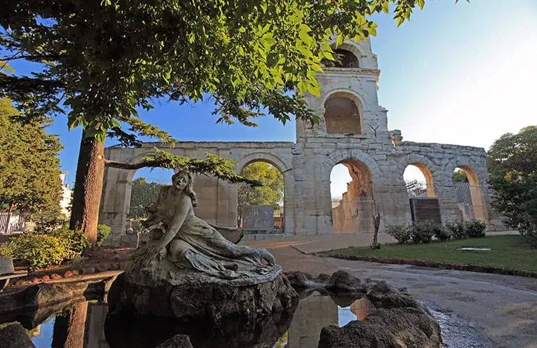 A fountain with the statue of a woman at the centre with a huge stone arched structure behind it in a park