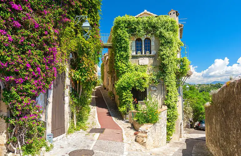 A pretty white stone building covered in green foliage with purple flowers with an alleyway leading up into the hills