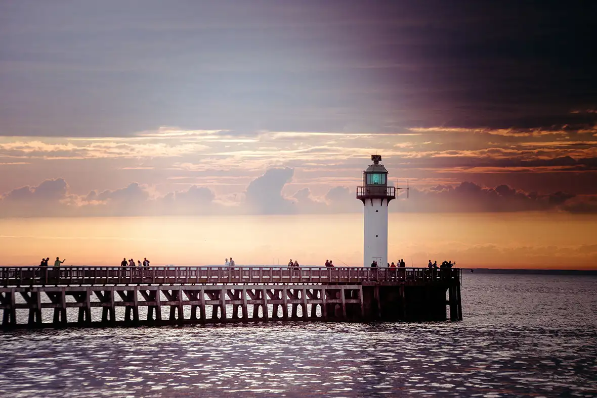 Lighthouse against a pink and orange sky at the end of a dark wood pier