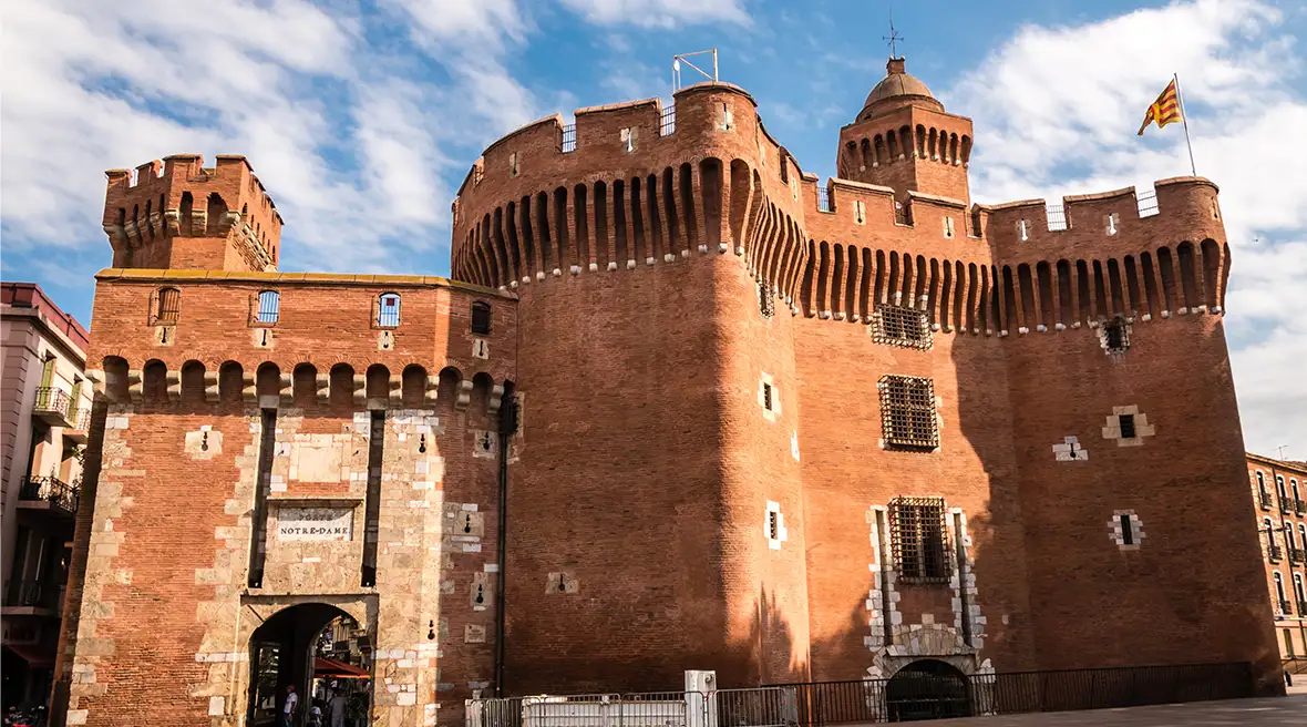Red brick building Les Castillet beneath a blue sky with white clouds, with a flag on one of the turrets