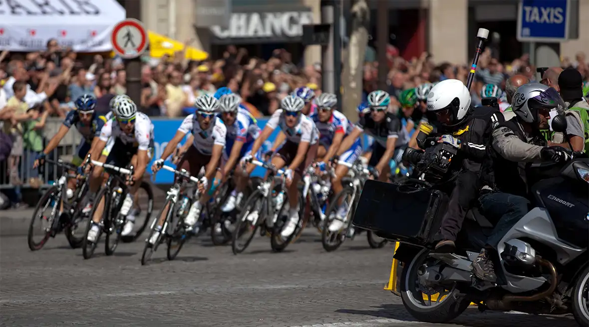 Cyclists in a road race swing round a corner in front of crowds behind crash barriers. Safety motorcyclist leads the way