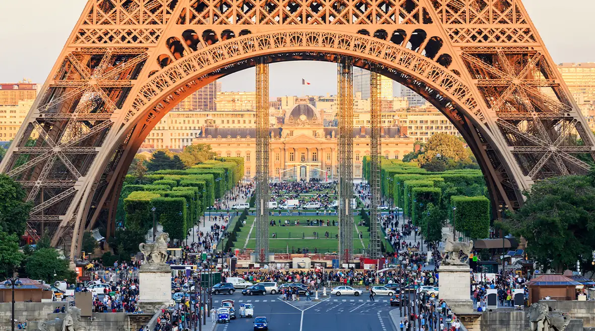 The base of the Eiffel Tower, underneath it a wide boulevard and garden with lots of crowds