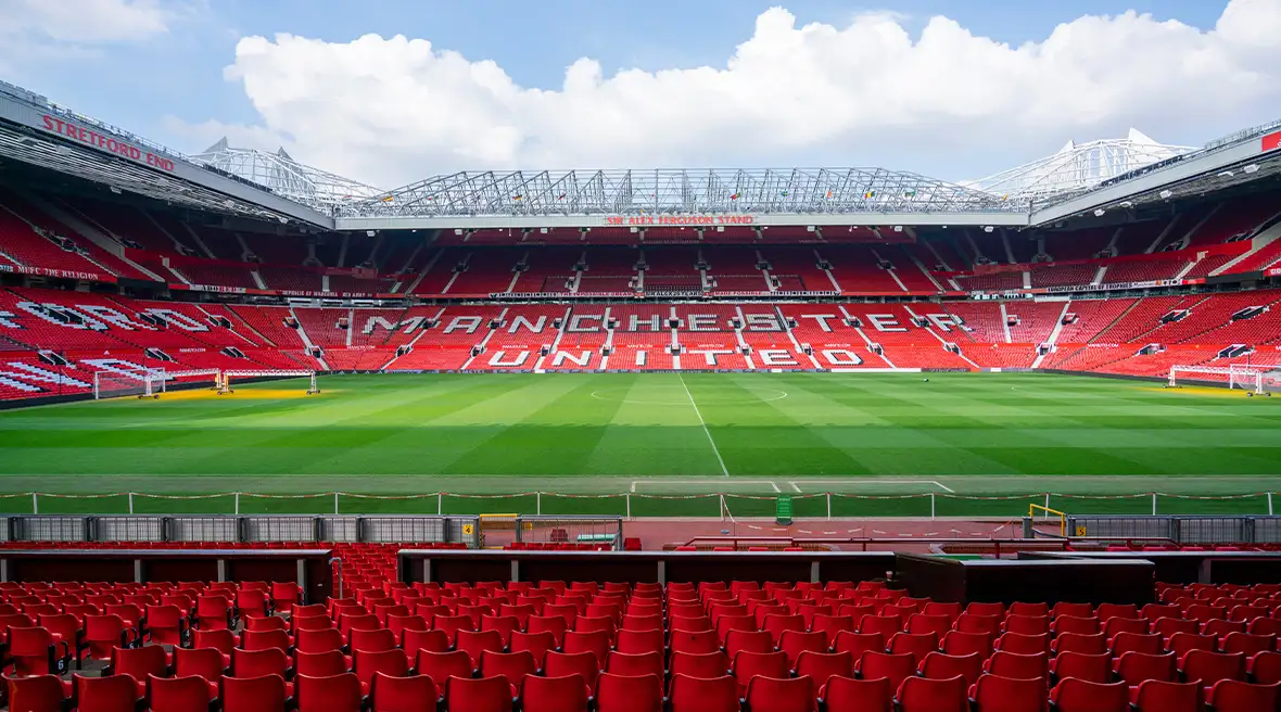 Vue de l’intérieur du stade Old Trafford avec sièges rouges en premier plan, pelouse et gradins avec l’inscription Manchester United en lettres blanches géantes