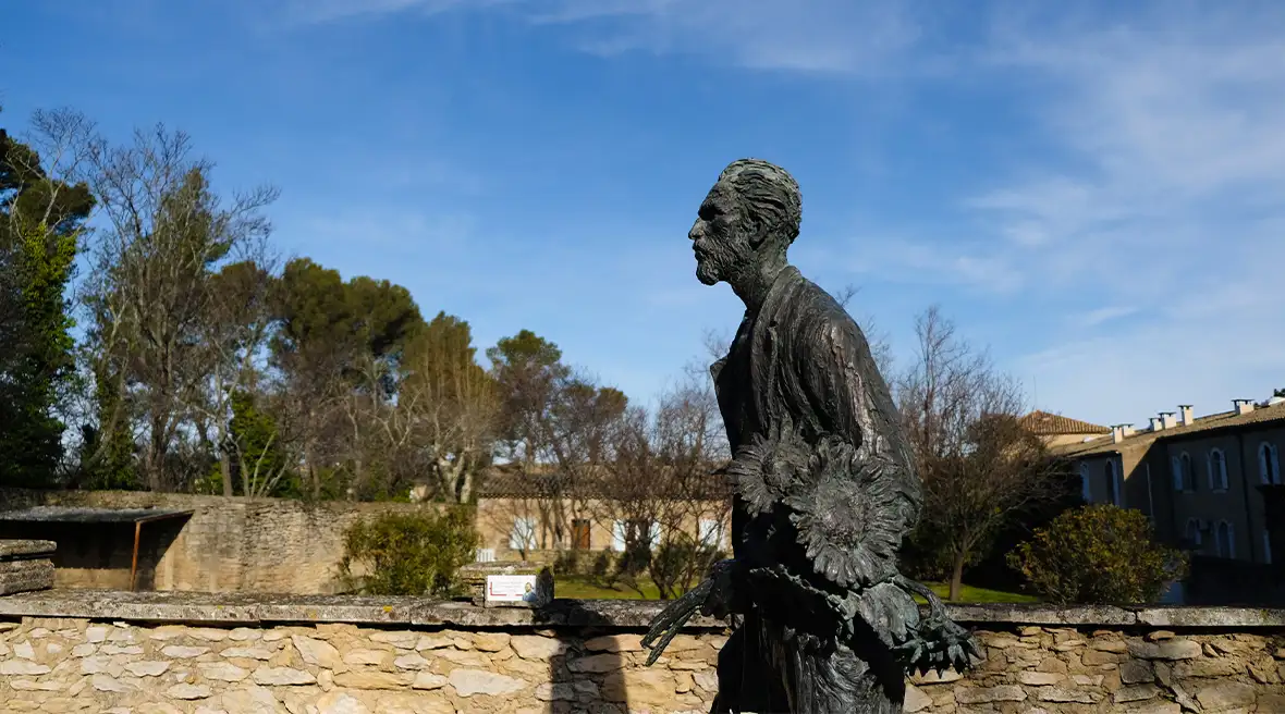 Black statue of a bearded man holding large sunflowers and paintbrushes, in front of a low wall on a summer’s day