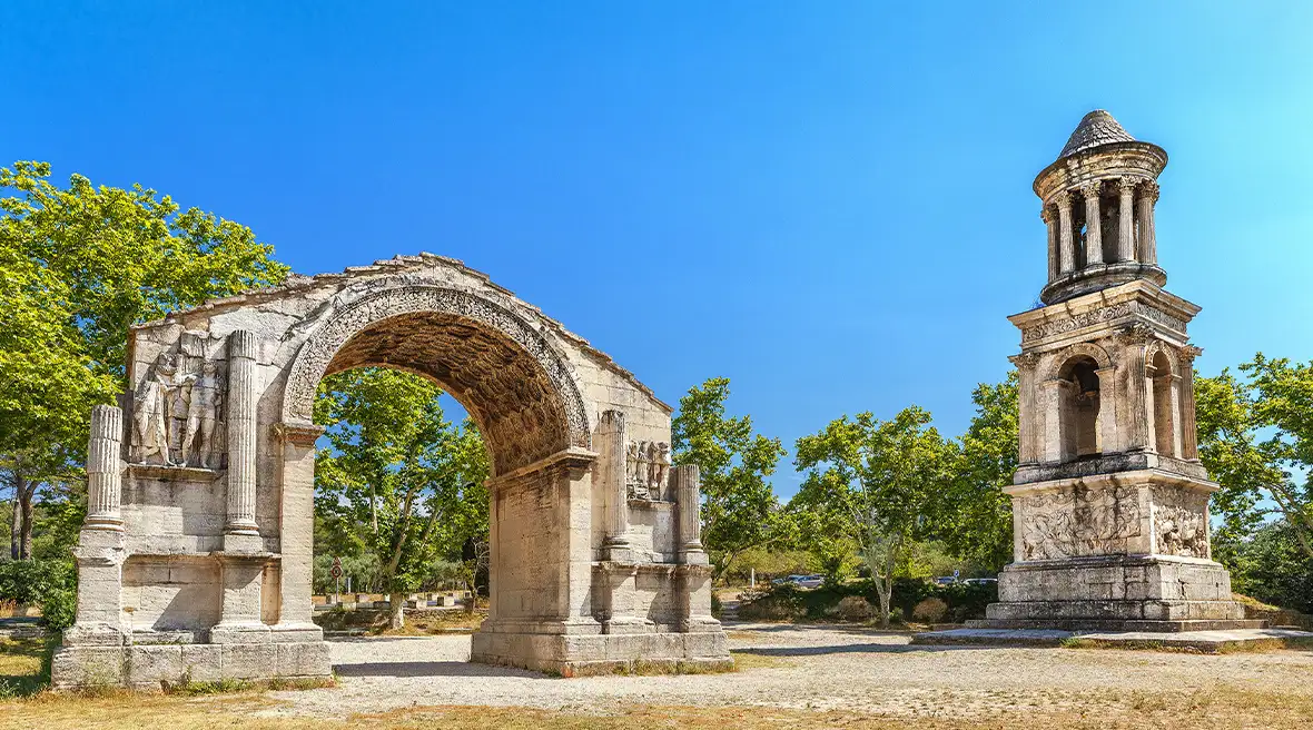 An ancient but well preserved stone arch and small tower stand close together, with trees behind them, with a cloudless blue sky behind