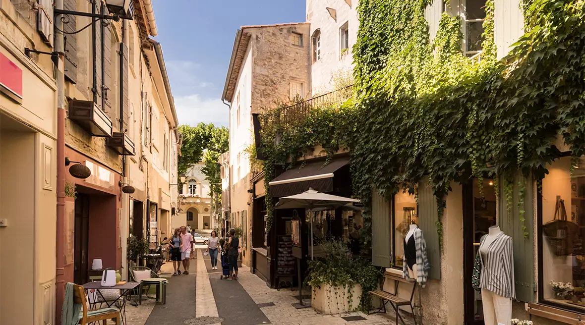 A narrow shopping street with an elegant clothes shop in the right foreground. Foliage covers most of the shop frontage