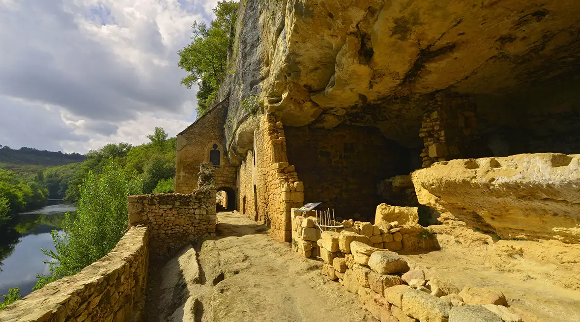A cave of sand-coloured stone with ruined walls by a river