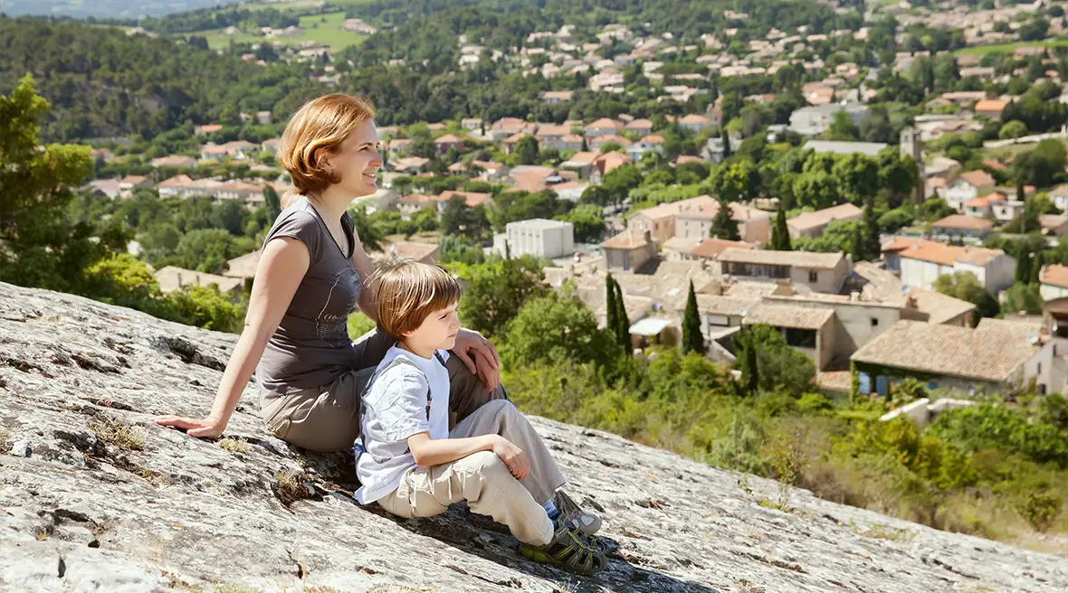 A mother and son staring across the view sitting on a sloping rock high above a town and greenery in the sunshine