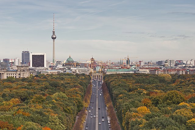 The Berlin TV Tower, with its iconic sphere, standing tall over city’s skyline. Lush green and autumnal trees line the road leading to the city.