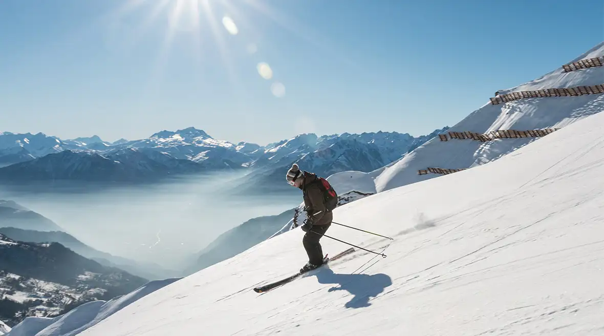 Someone skiing down a slope against a bright blue sky and a mountain range in the distance