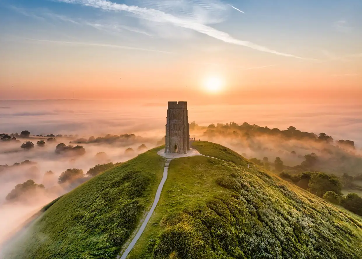 De heuvel Glastonbury Tor vanuit de lucht met St Michael’s Tower verlicht door de opgaande zon.