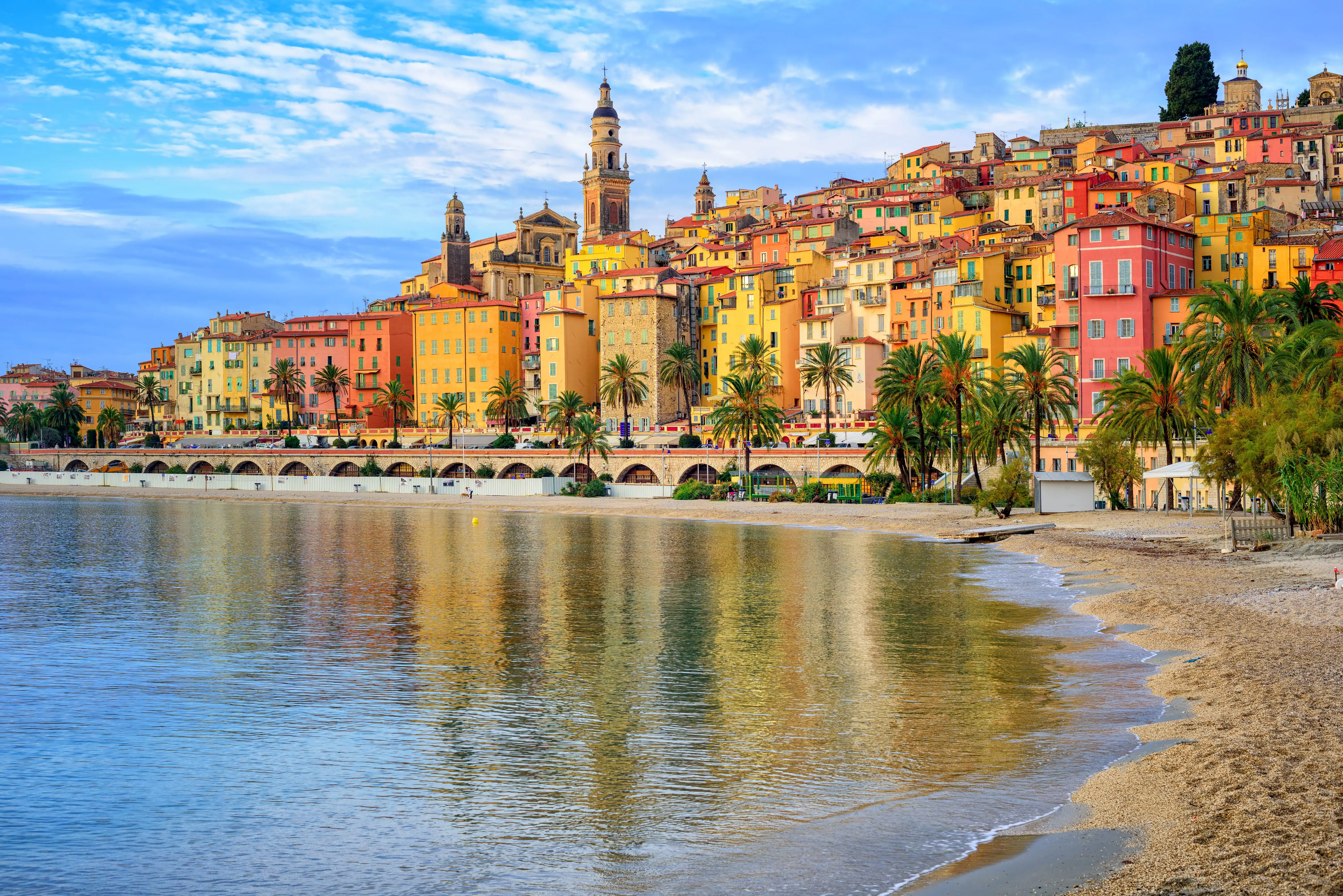 Pastel coloured buildings of a seafront town, with palm trees and a promenade, Menton, France